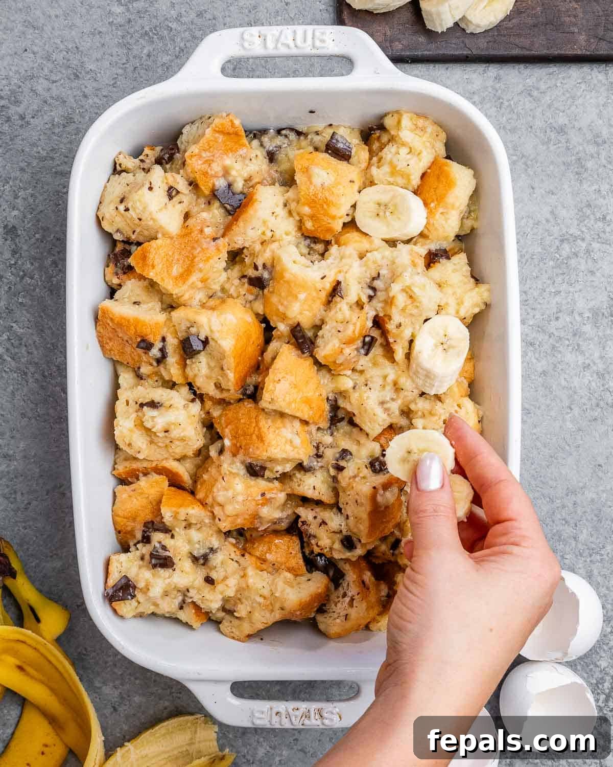 The banana bread pudding mixture being transferred into a prepared baking dish, with a hand arranging fresh sliced bananas on top before baking.