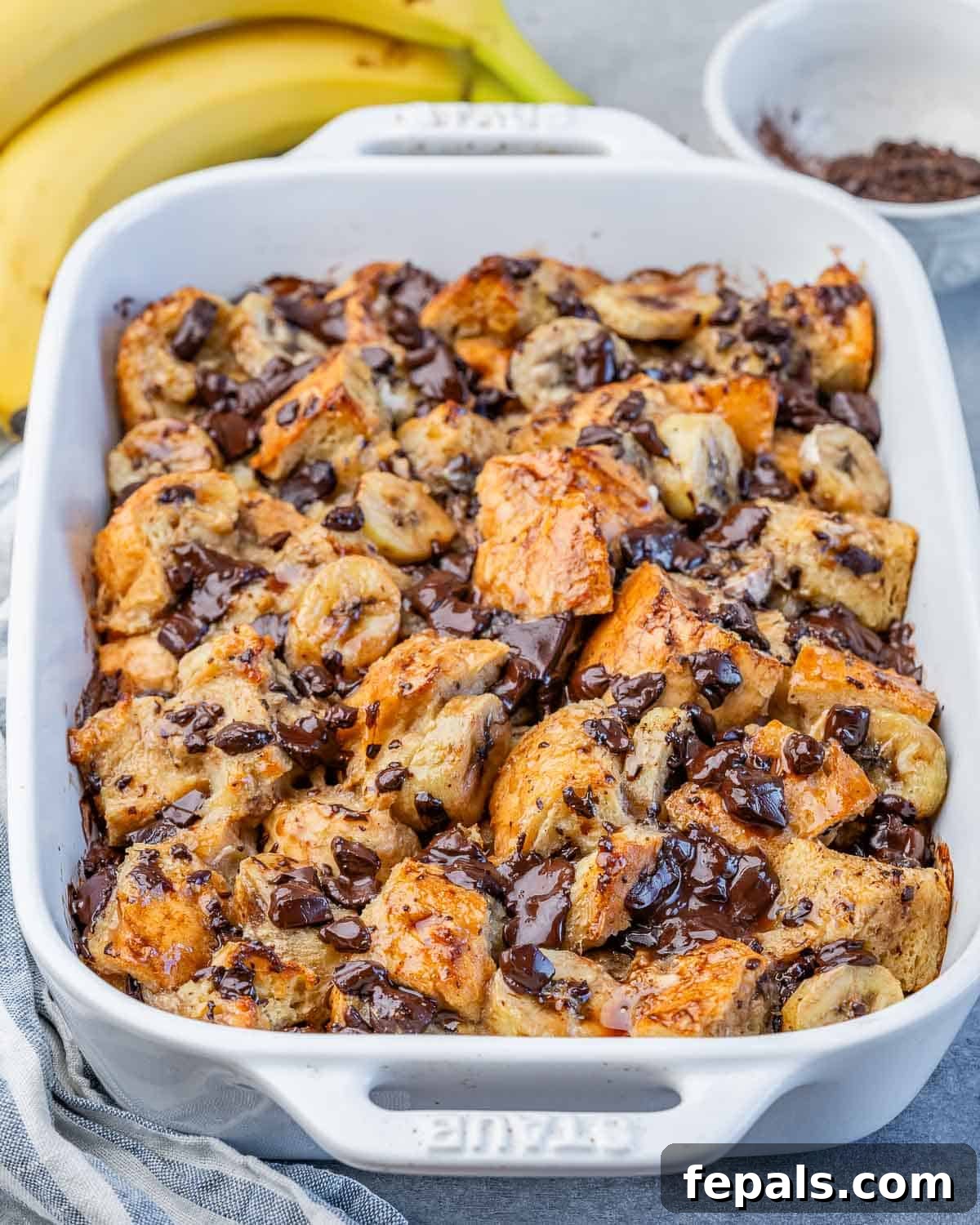A large white baking dish filled with freshly baked banana bread pudding, beautifully golden with visible melted chocolate and banana slices on top.