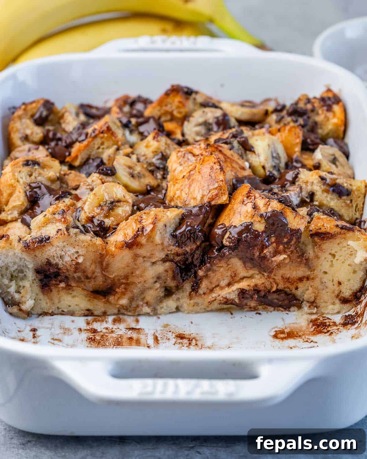 A close-up shot of a half-eaten banana bread pudding in a white baking dish, showing its rich, custardy texture and pieces of chocolate and banana.