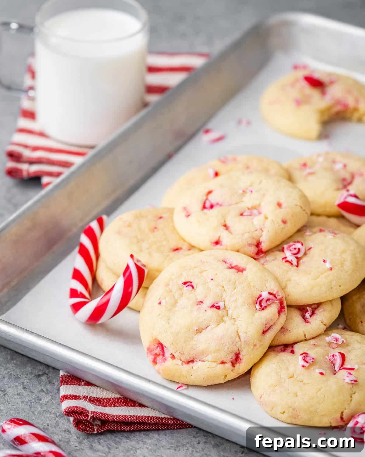 White sugar cookies adorned with festive peppermint pieces on a baking sheet, with a candy cane on the side, creating a perfect holiday scene.