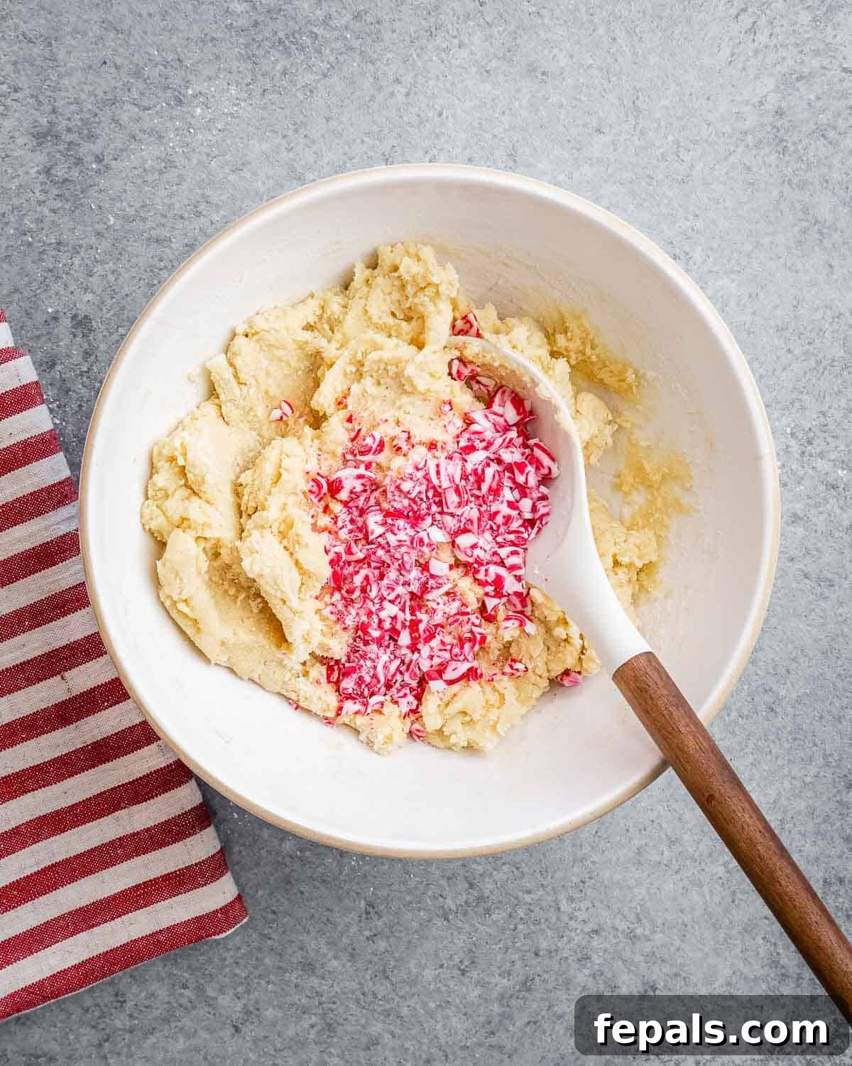 Brightly crushed candy cane pieces being gently folded into the prepared cookie dough, adding a festive crunch and burst of peppermint flavor.