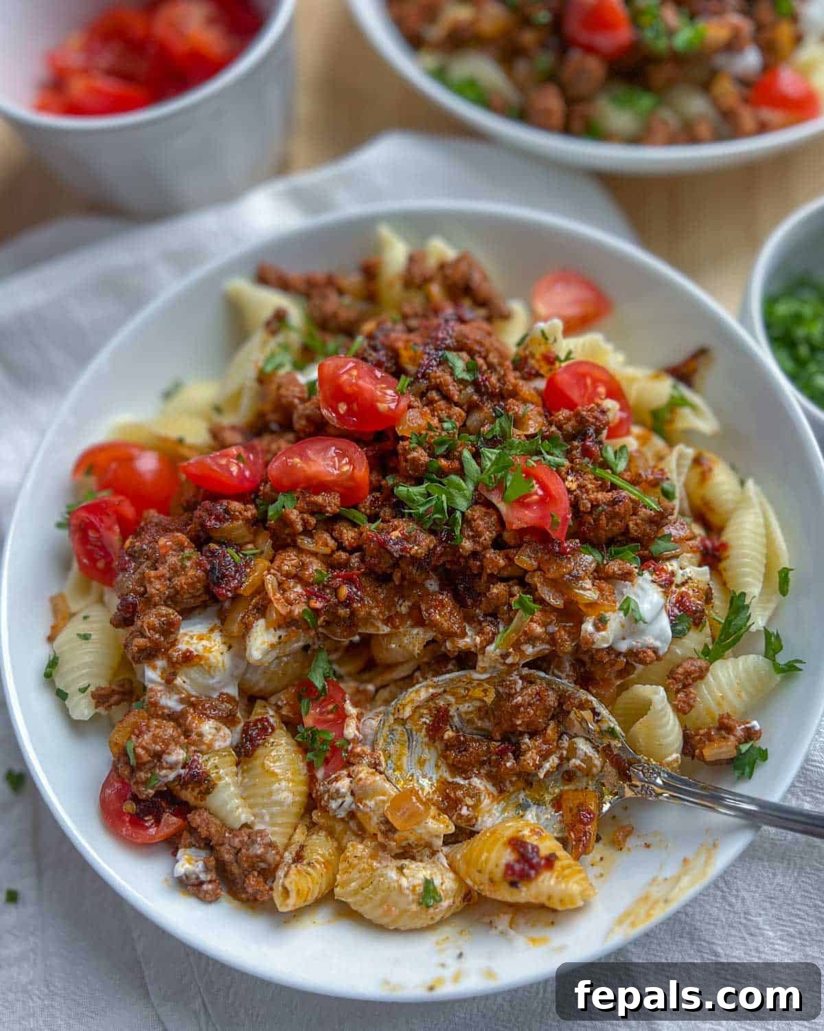 A spoon gracefully lifts a portion of Turkish pasta, revealing the rich meat sauce, creamy yogurt, and vibrant garnish of chopped tomatoes and parsley.