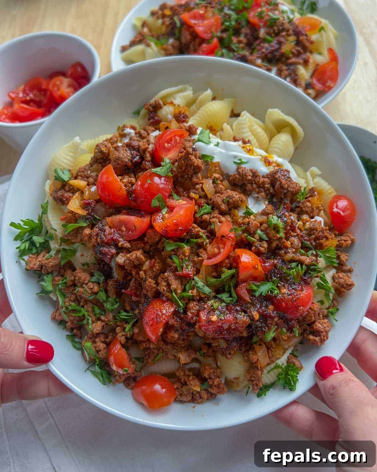 A close-up of a hand holding a plate of the vibrant Turkish pasta, perfectly garnished with fresh chopped tomatoes and parsley, ready to be enjoyed.