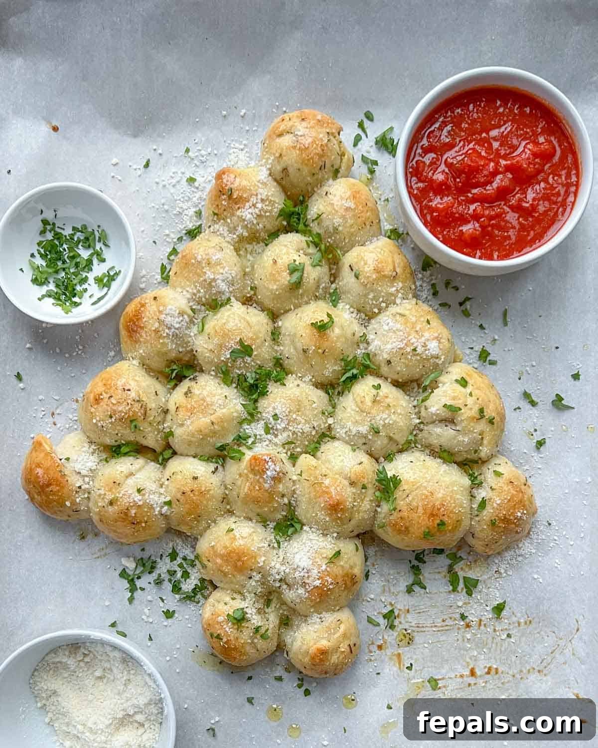 A festive Christmas tree-shaped pull-apart cheese bread, golden brown and garnished with herbs, served with a bowl of rich marinara sauce and grated mozzarella.