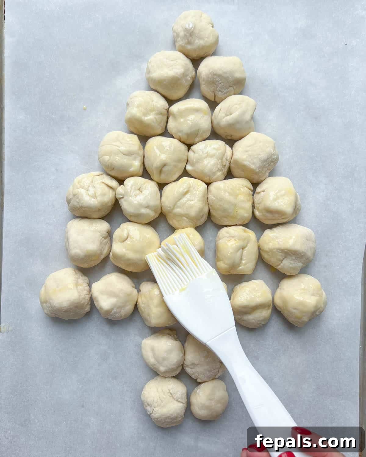 A whisked egg wash being delicately brushed over the unbaked, Christmas tree-shaped arrangement of cheese dough balls.