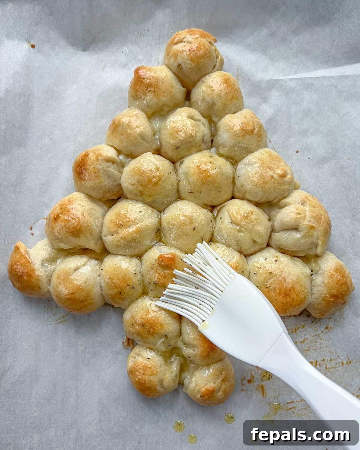 A savory herb-infused oil being brushed over the freshly baked, golden-brown cheese balls of the Christmas tree pull-apart bread.
