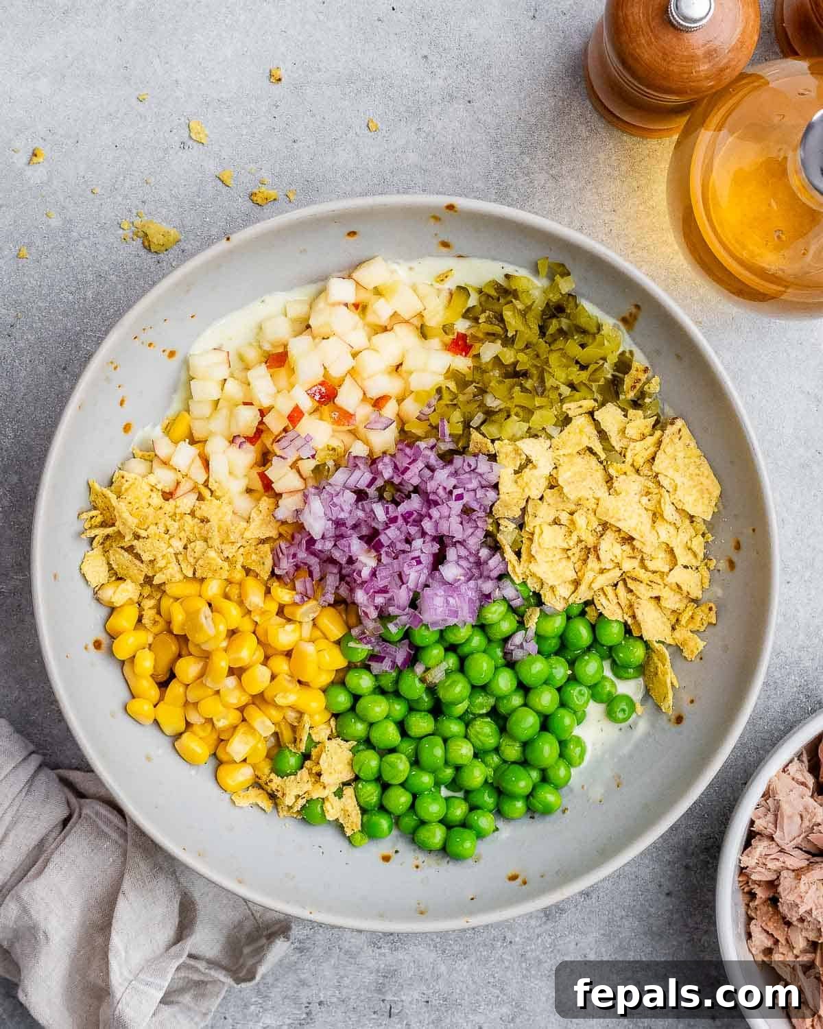 The McConaughey Tuna Salad That Broke the Internet 6 A bowl filled with a colorful array of chopped ingredients, including thawed peas, corn kernels, red onion, apple, and pickles, layered over the dressing.