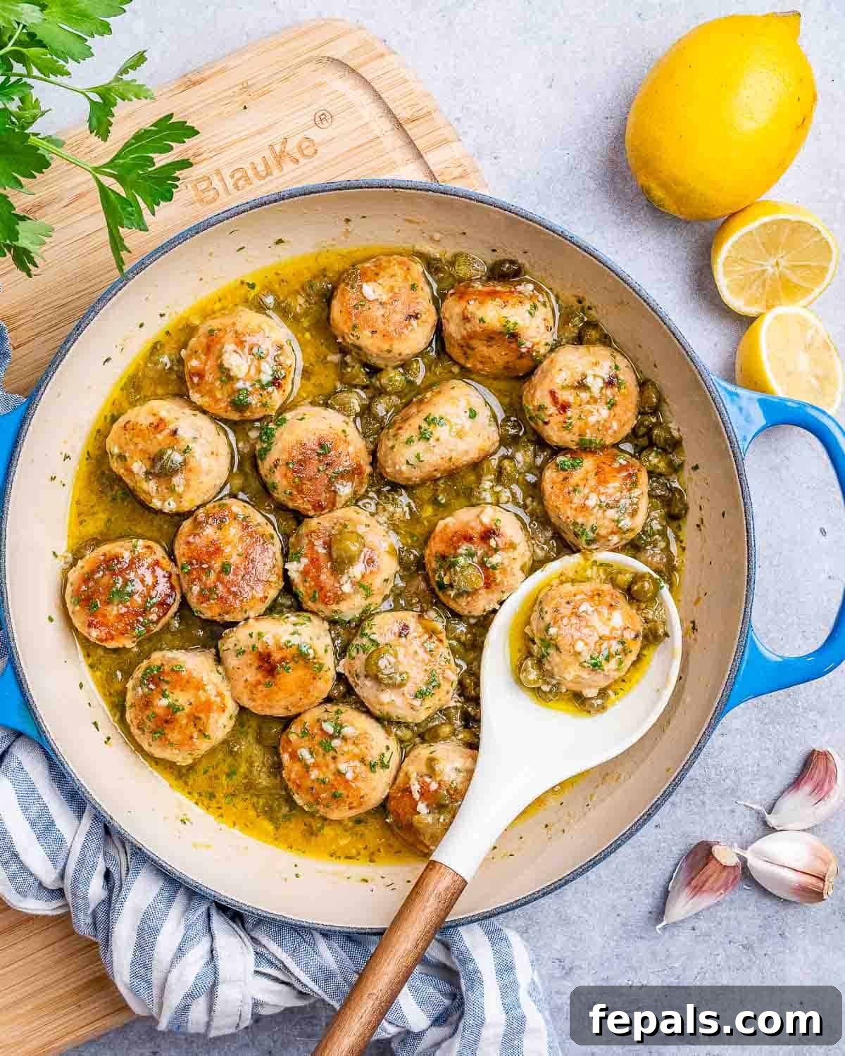A white spoon holding a meatball in a pot filled with chicken meatballs in a rich, lemon-caper sauce.