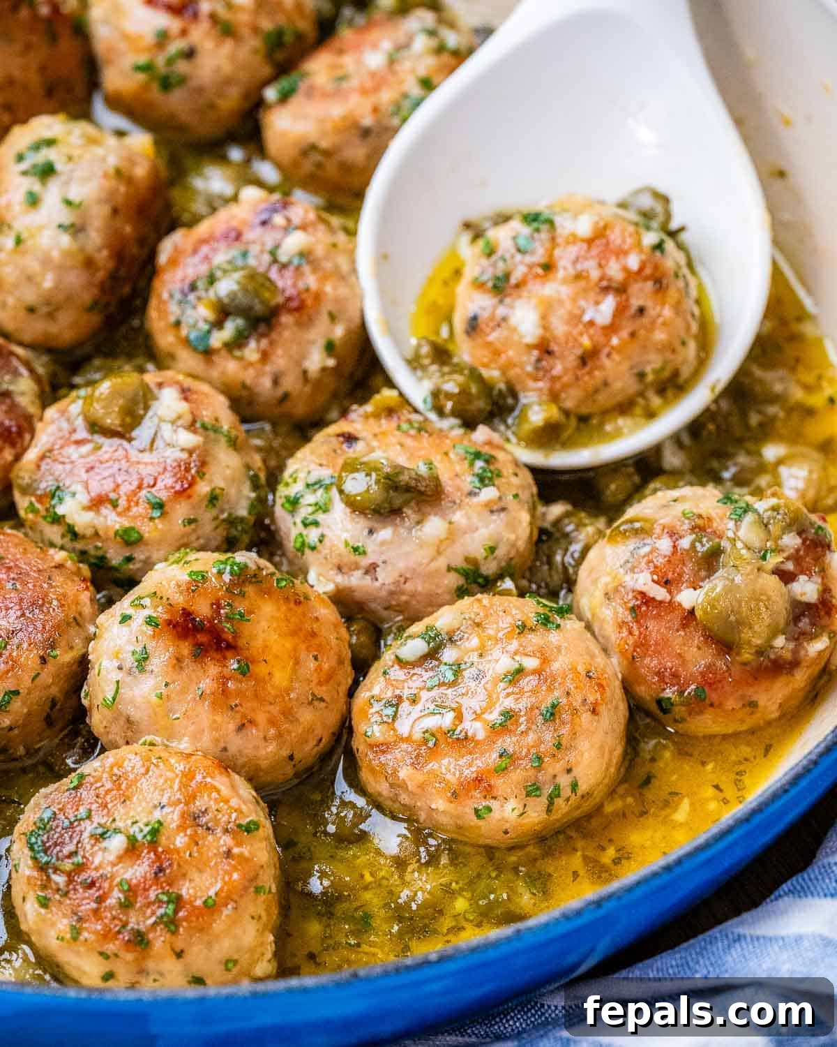 A white spoon serving a golden-brown chicken meatball from a pot filled with rich, lemony sauce and more meatballs.
