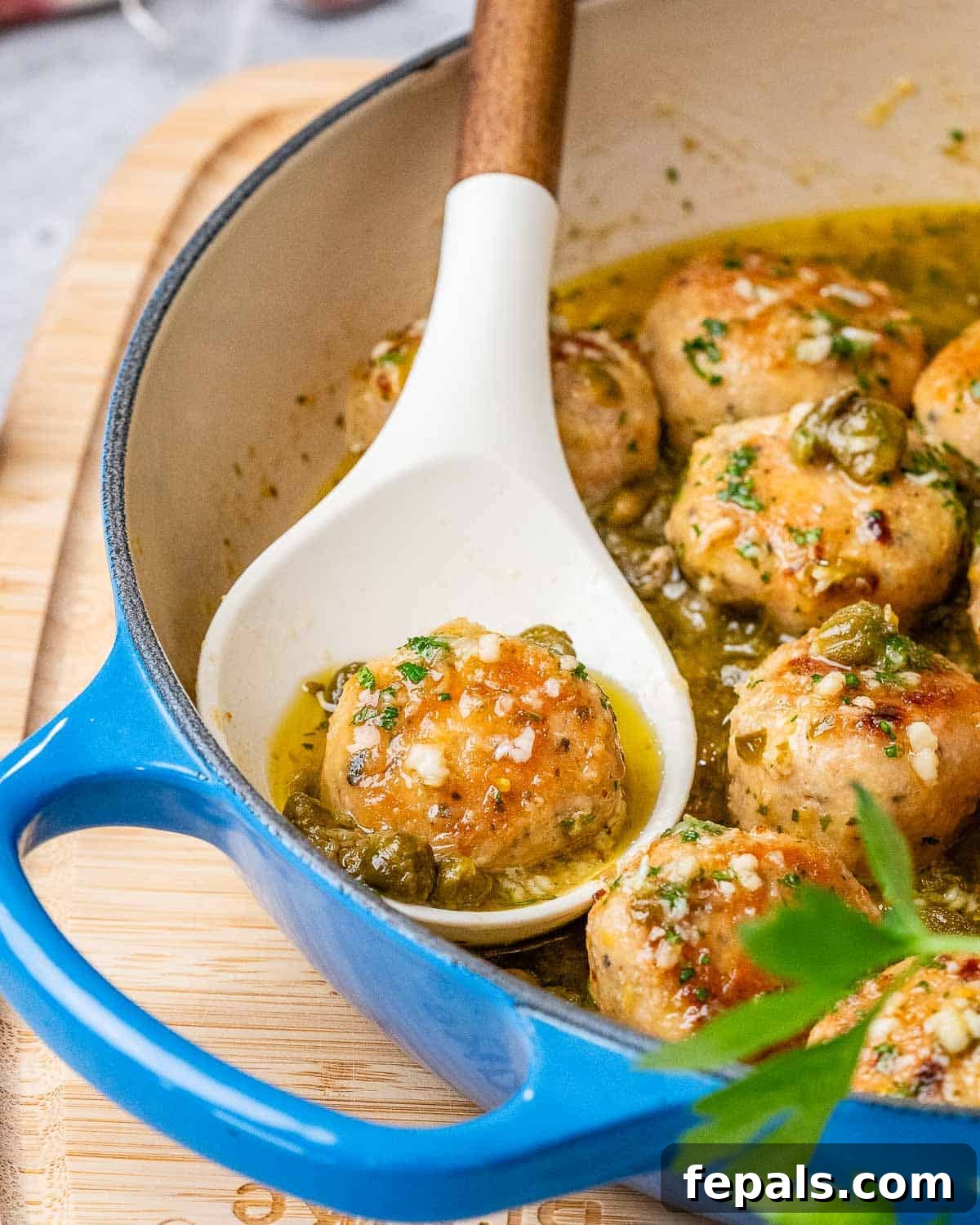 A close-up of golden-brown chicken piccata meatballs in a blue skillet, with one meatball on a white spoon.
