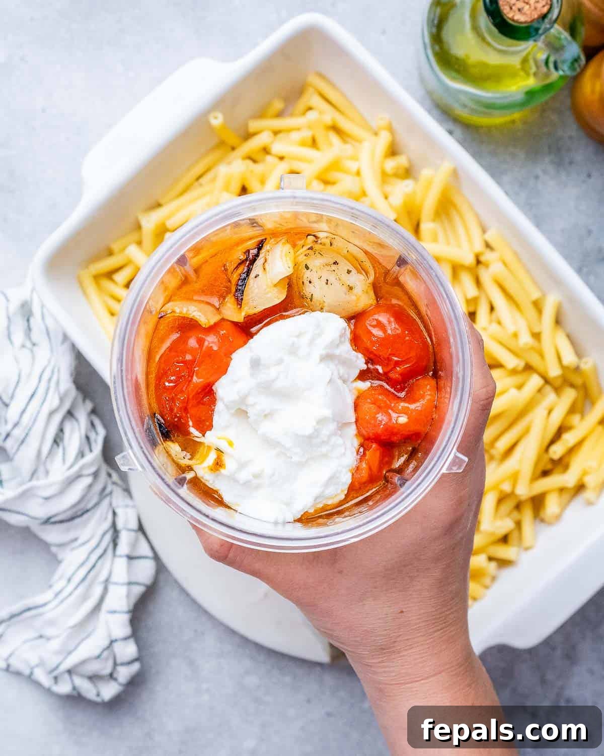 Ultimate Cheesy Baked Ziti 7 A hand holding a plastic container, adding creamy ricotta cheese to the roasted vegetable mixture in a casserole dish.