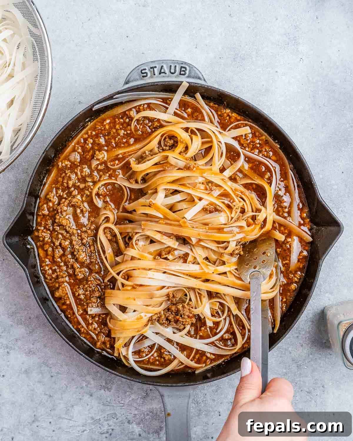 Cooked rice noodles being tossed with ground beef and Mongolian sauce using kitchen tongs in a skillet.