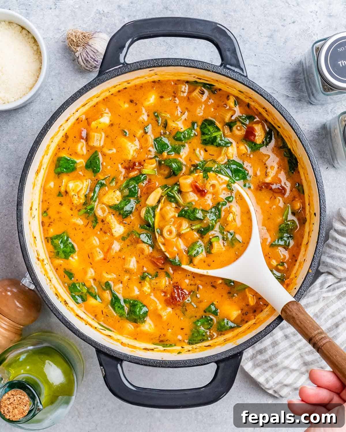 A white spoon holding a scoop of soup above a pot on a gray surface, showcasing the rich, creamy texture and visible pieces of chicken and sun-dried tomatoes.