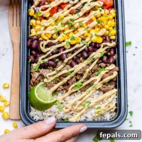 A hand holding a black meal prep bowl filled with a savory beef burrito mix, featuring wholesome brown rice, black beans, sweet corn, seasoned ground beef, and fresh salsa, all crowned with a creamy sauce.