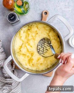 Homestyle Beef Shepherd's Delight 15 A hand using a potato masher to mash potatoes in a steel pot.