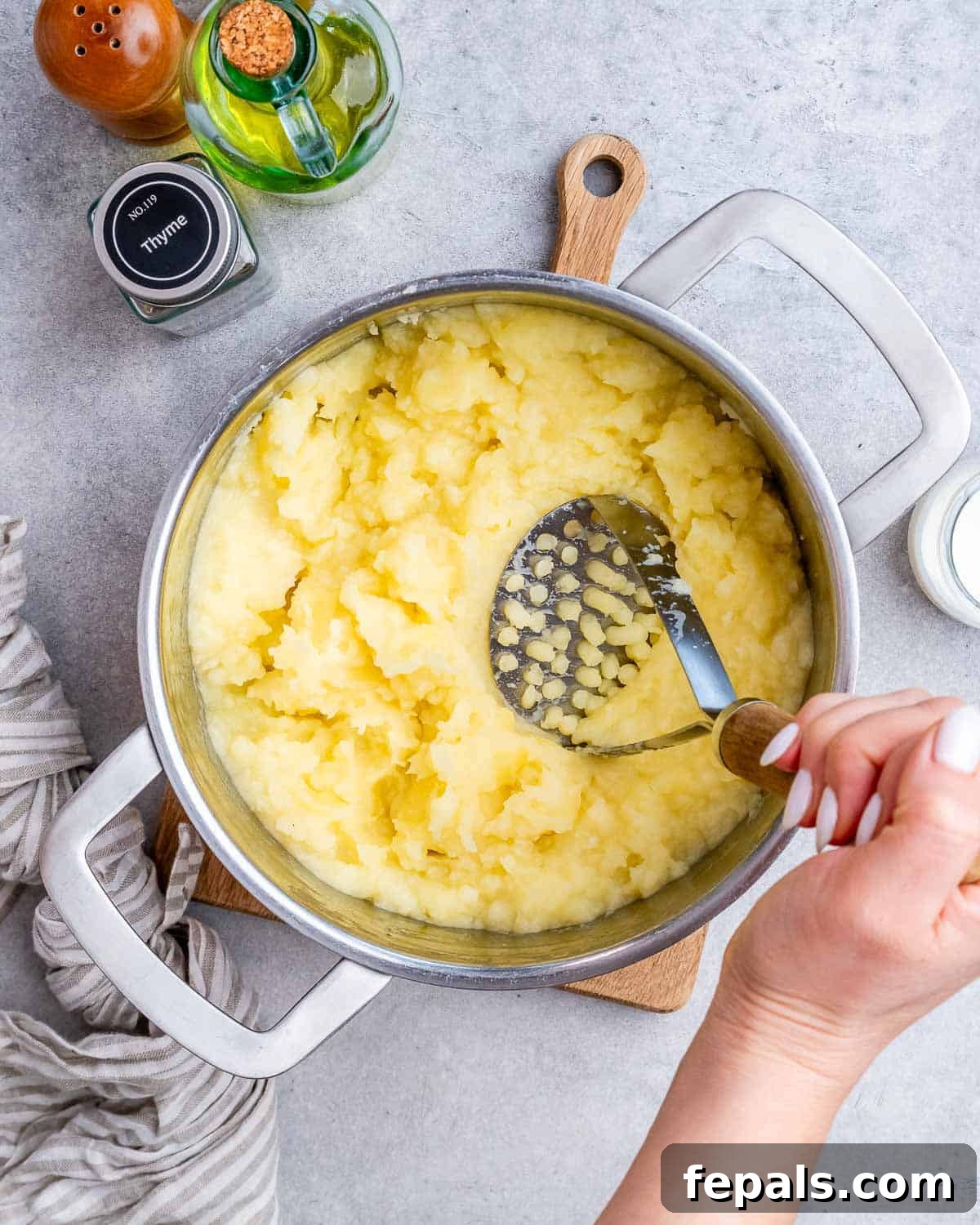 Homestyle Beef Shepherd's Delight 7 A hand using a potato masher to mash potatoes in a steel pot.