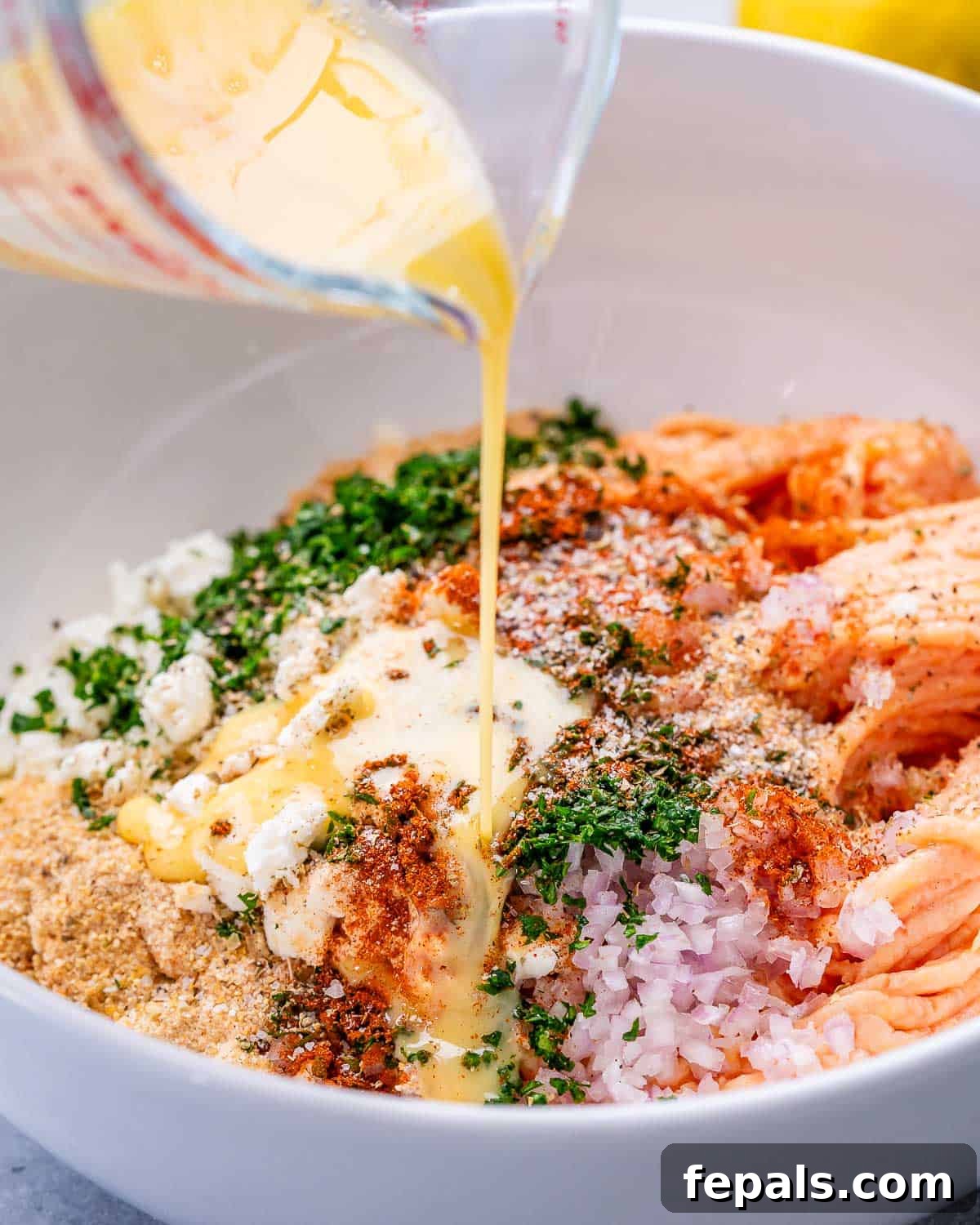 A clear measuring cup pouring whisked egg mixture, infused with fresh parsley and spices, over the meatball ingredients in a white bowl.