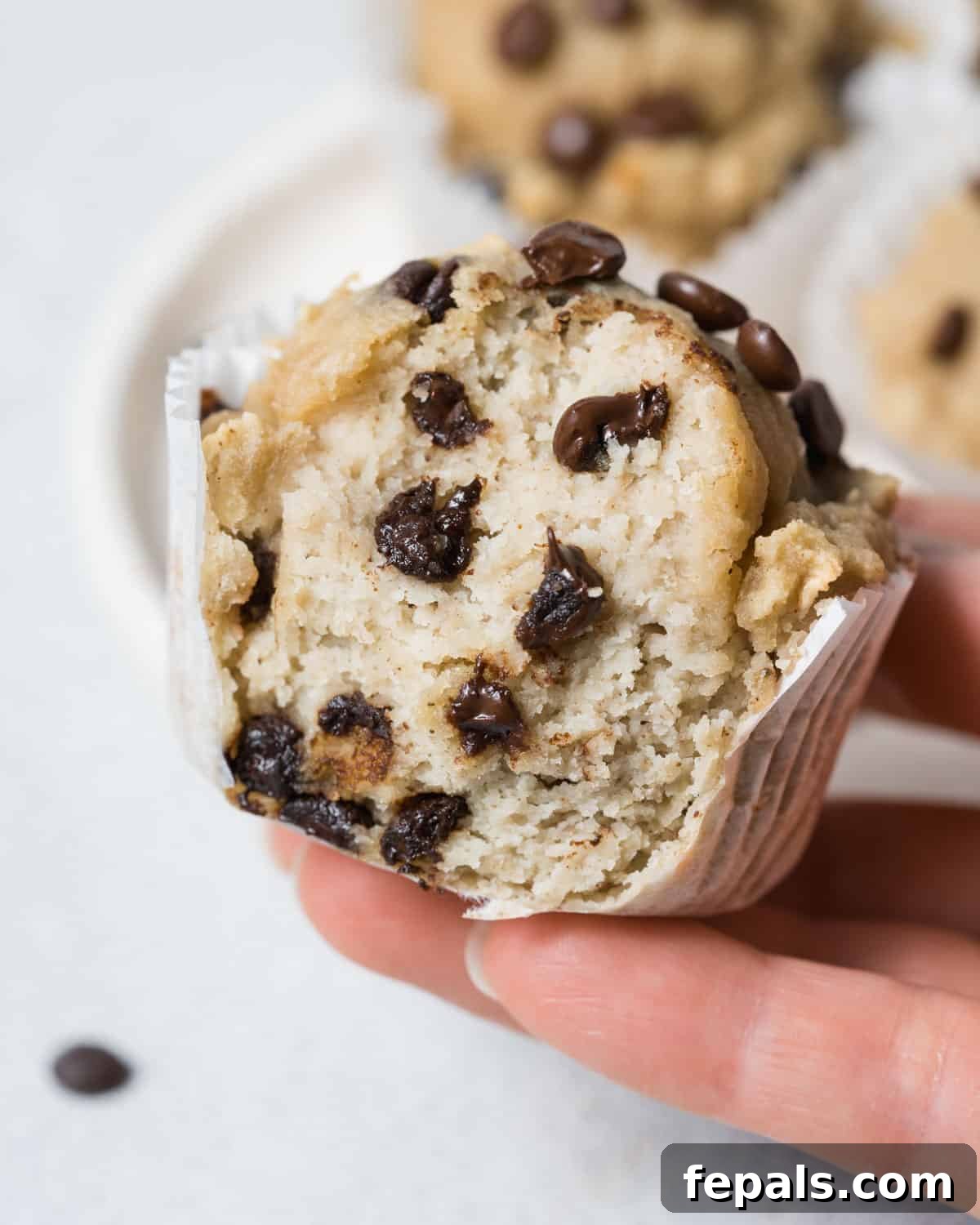 A hand holding a perfectly baked almond flour banana muffin, cut in half to reveal its moist interior and chocolate chips.