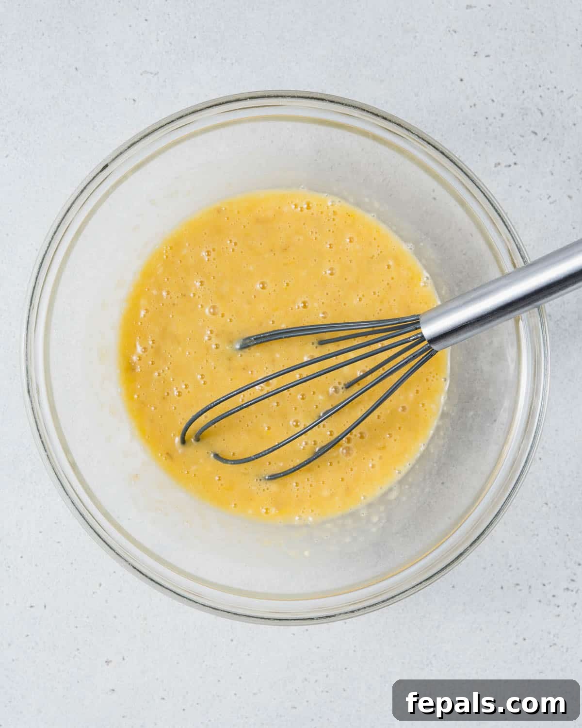 Mashed ripe bananas, whisked eggs, maple syrup, and vanilla extract combined in a large mixing bowl, creating a smooth wet mixture.
