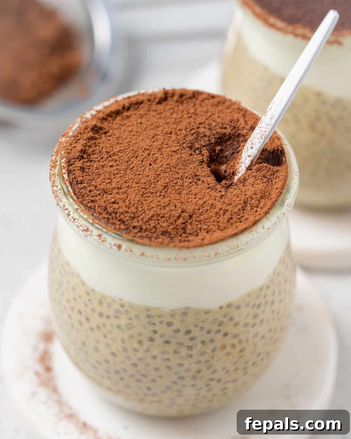 A spoon in a jar of tiramisu chia pudding layered with yogurt and cocoa powder, ready to be enjoyed as a healthy breakfast or dessert.