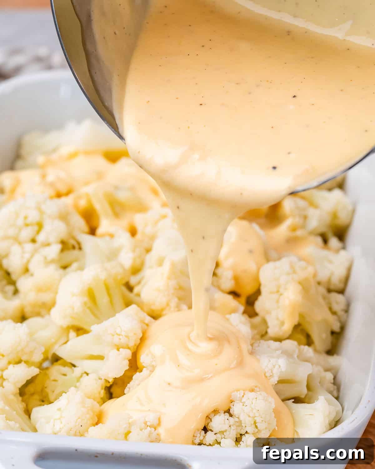 Pouring cheese sauce over chopped cauliflower in a baking dish.