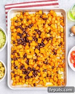 Roasted diced sweet potatoes and black beans spread on a baking sheet, surrounded by small bowls of ingredients.