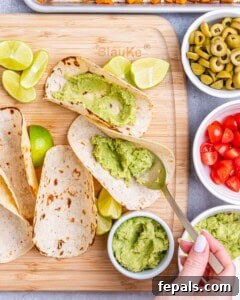 Tortillas being filled with guacamole on a wooden board, surrounded by small bowls of tomatoes, olives, and lime wedges.