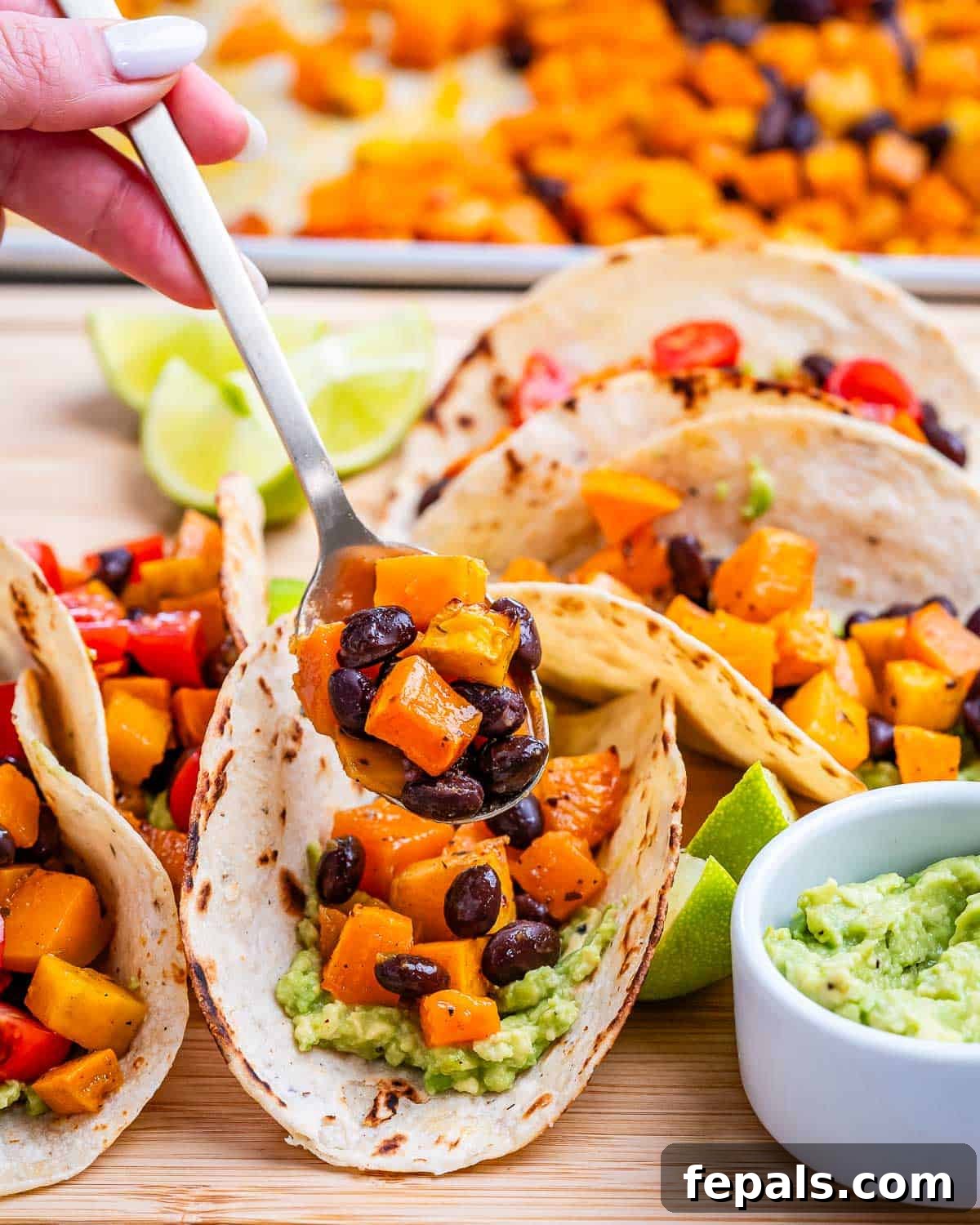 Close up view of tacos being assembled, with a spoon adding filling into a tortilla, surrounded by guacamole and lime.