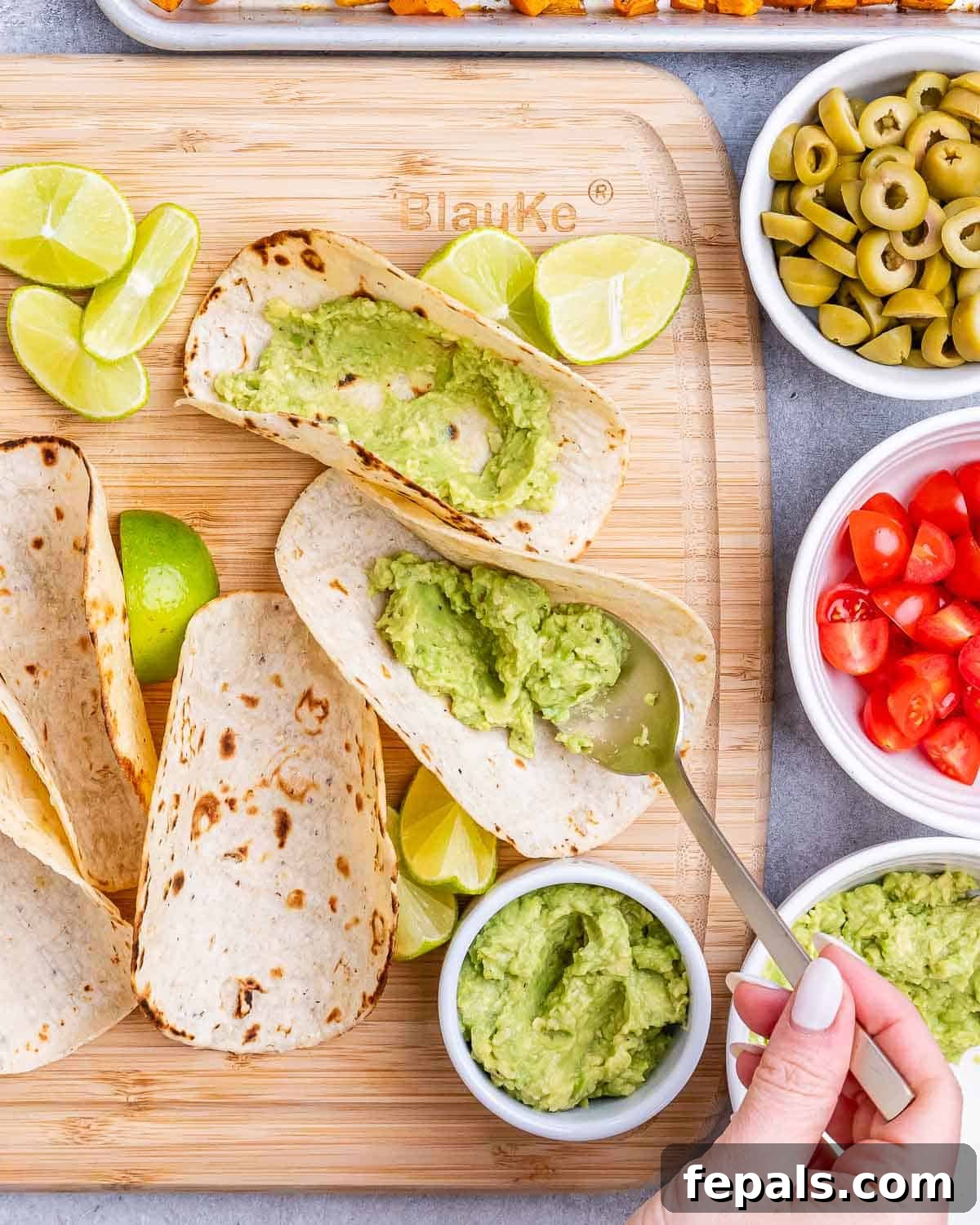 Warm corn tortillas being spread with guacamole on a wooden serving board, ready for filling.