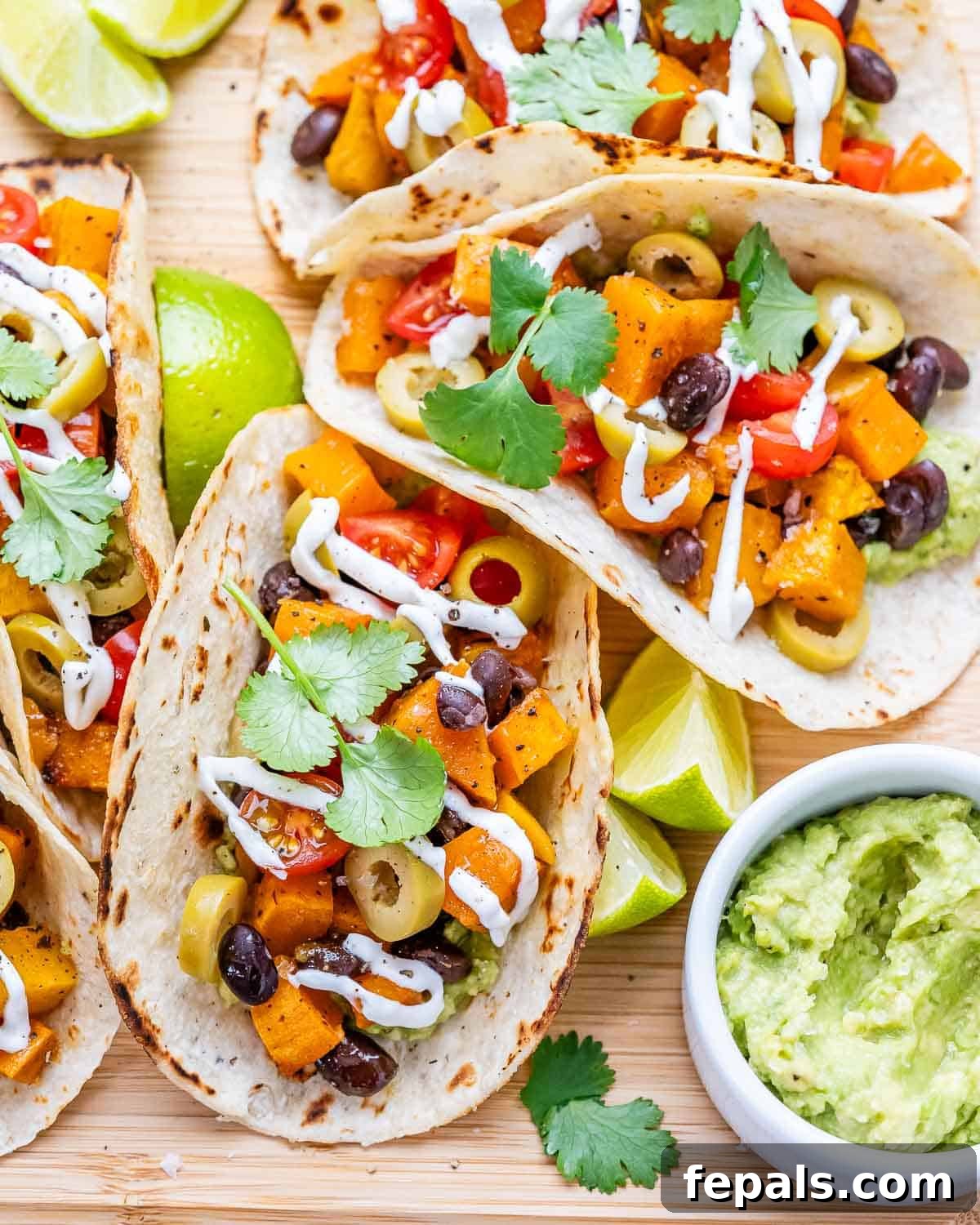 Three fully assembled Black Bean Sweet Potato Tacos on a wooden board, topped with tomatoes, olives, and cilantro, with lime wedges and guacamole on the side.
