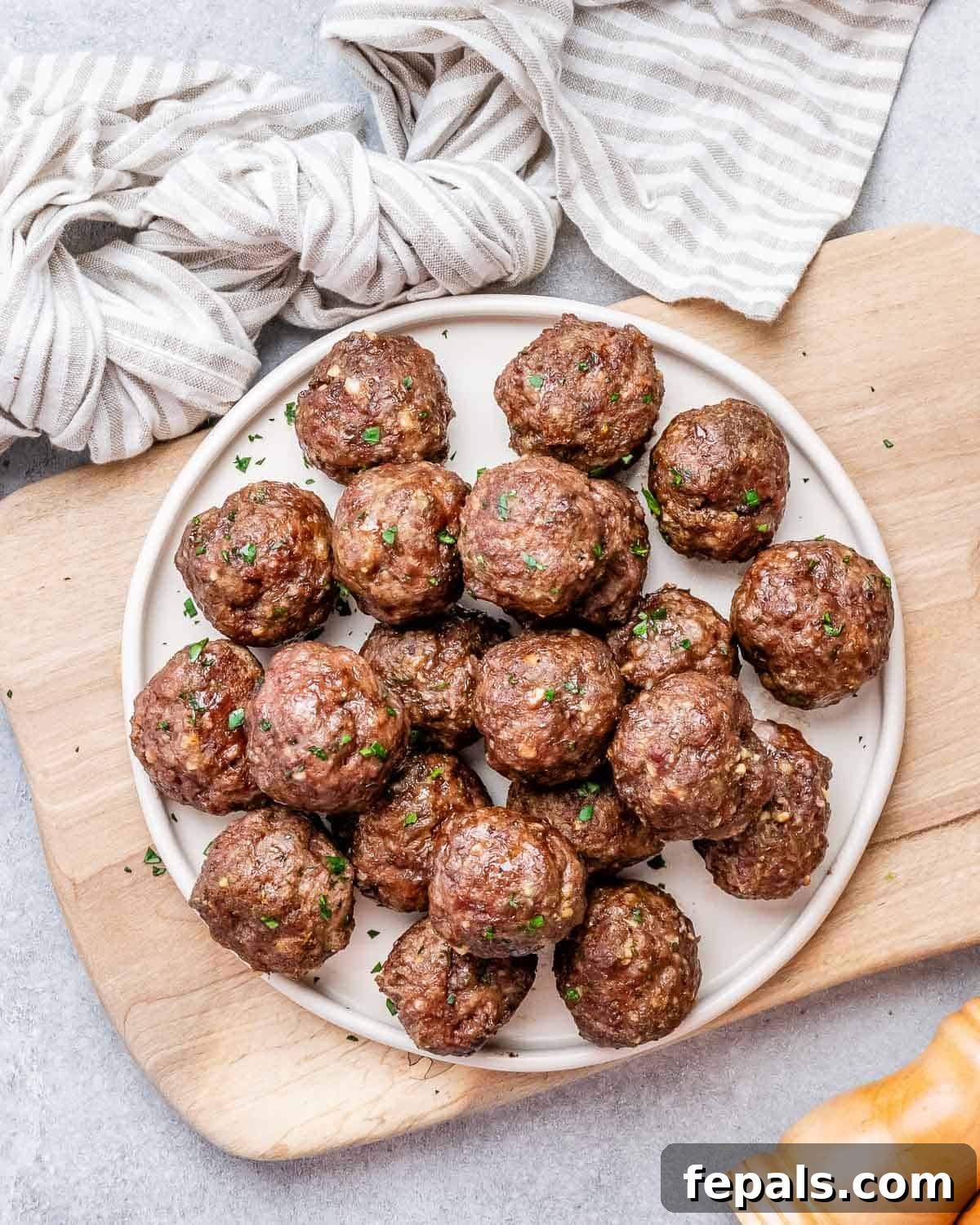 Golden Air Fryer Meatballs 2 A white round plate filled with perfectly cooked, golden-brown air fryer meatballs, resting on a rustic wooden cutting board, garnished with fresh herbs.