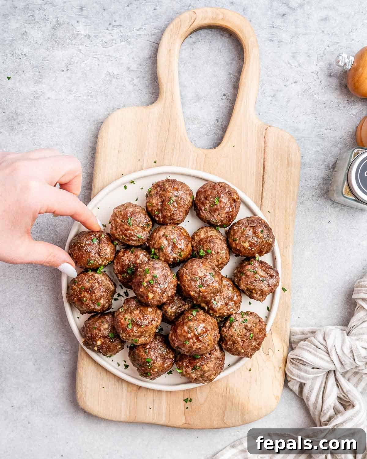 Golden Air Fryer Meatballs 9 A hand reaching for a delicious air fryer meatball from a white round plate, filled with perfectly cooked meatballs on a wooden cutting board.