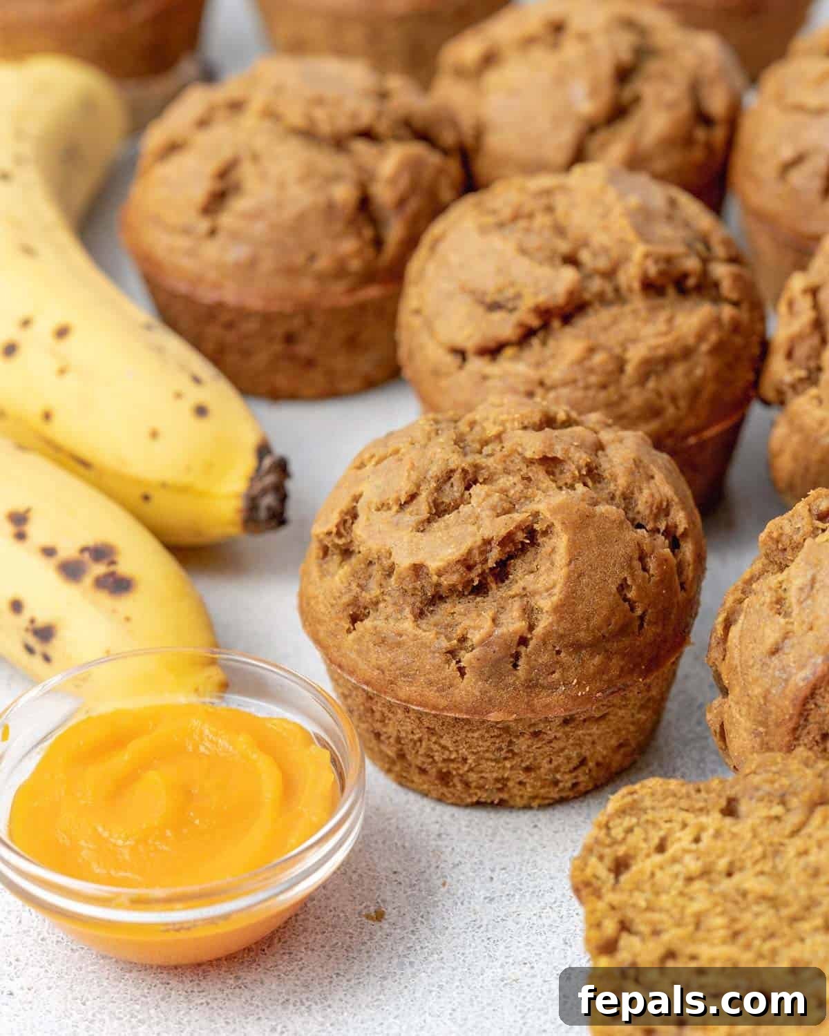 Pumpkin banana muffins on a white surface next to a bowl of pumpkin puree and some bananas. Golden-brown muffins are perfectly domed, with a few fresh bananas and a small white bowl of orange pumpkin puree visible in the background, hinting at the key ingredients.