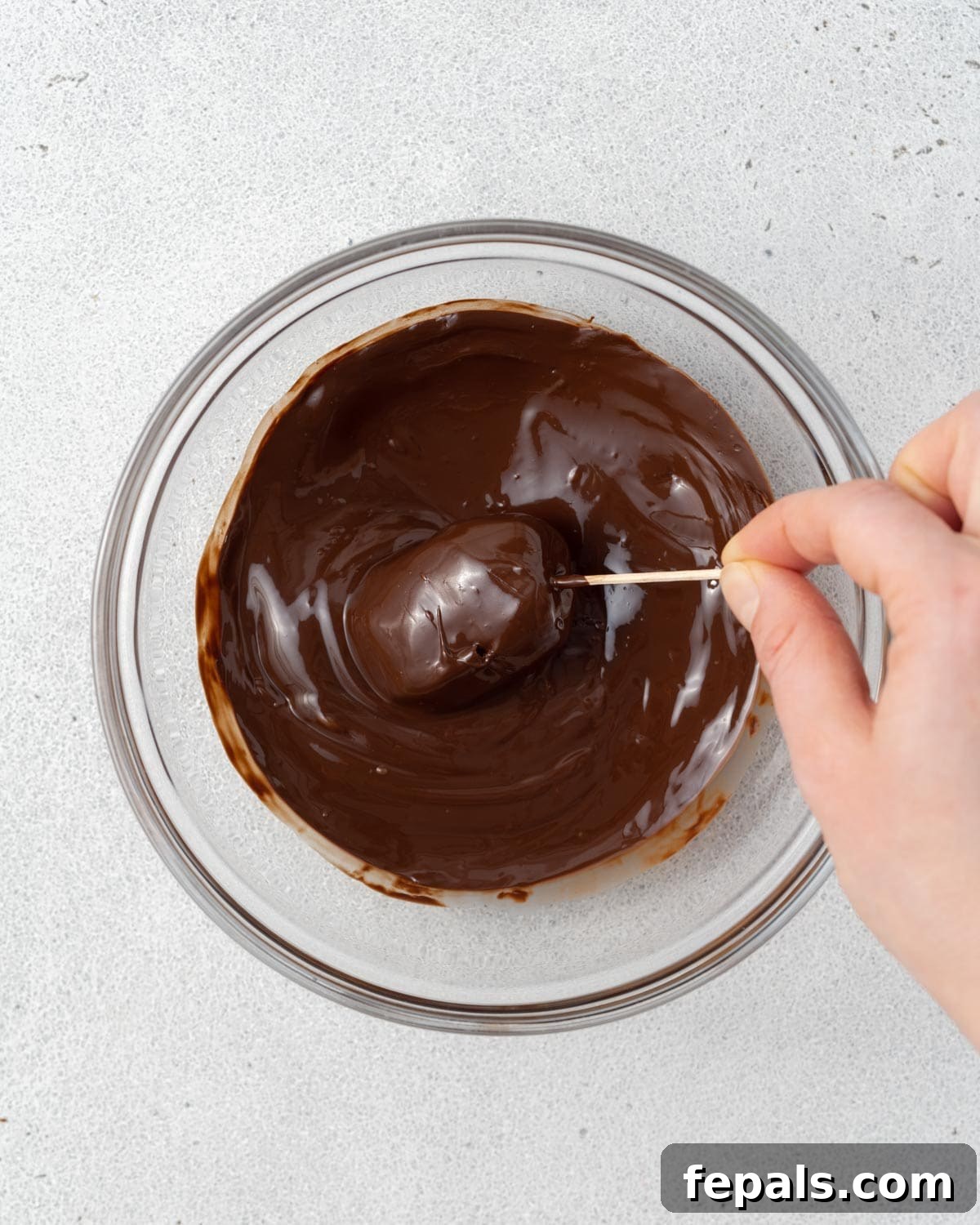 A hand using a toothpick to pierce the date and bring it out of the melted chocolate.