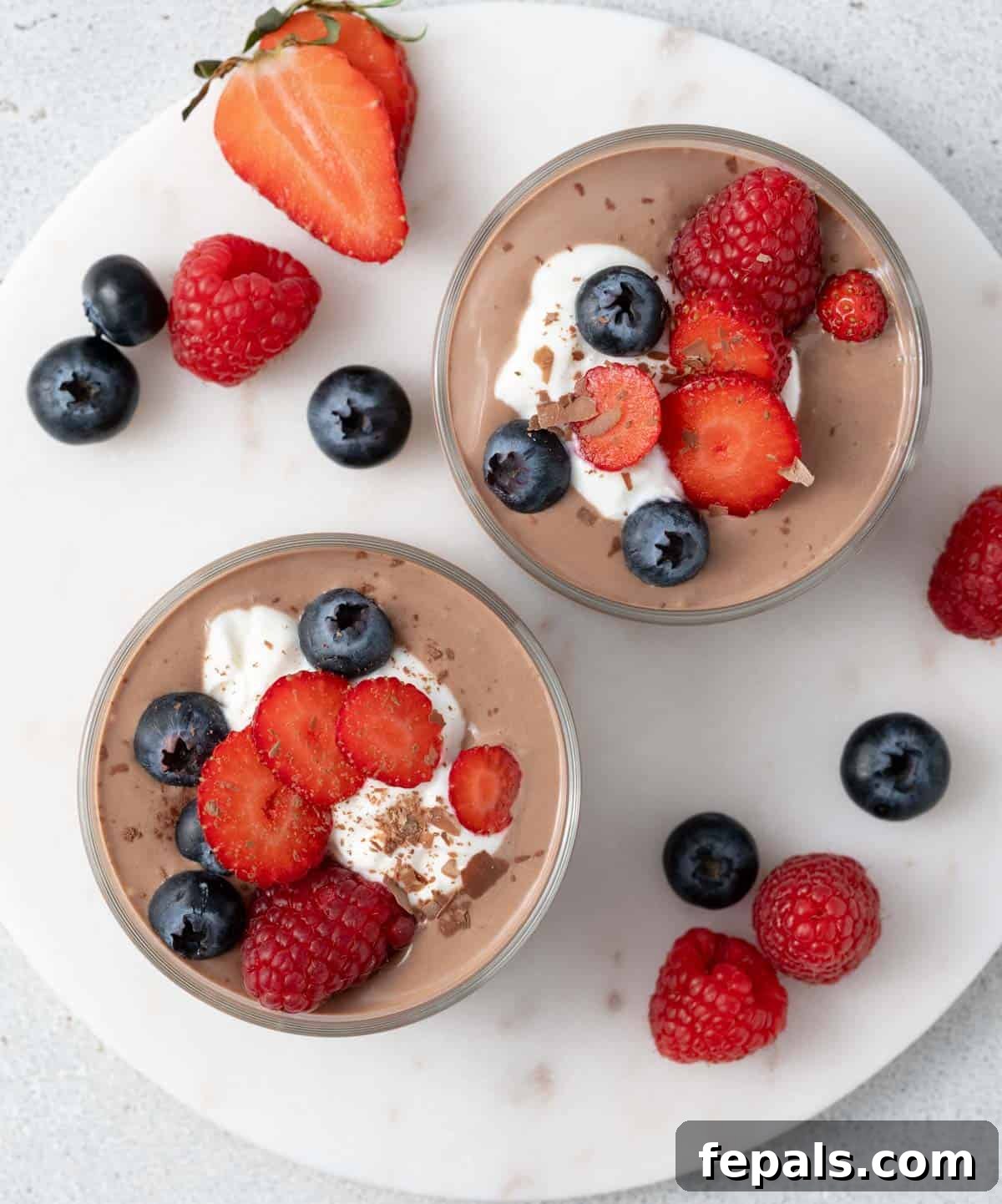 Top view of chocolate mousse topped with whipped cottage cheese, sliced strawberries, raspberries, and blueberries in glass cups on a white surface.