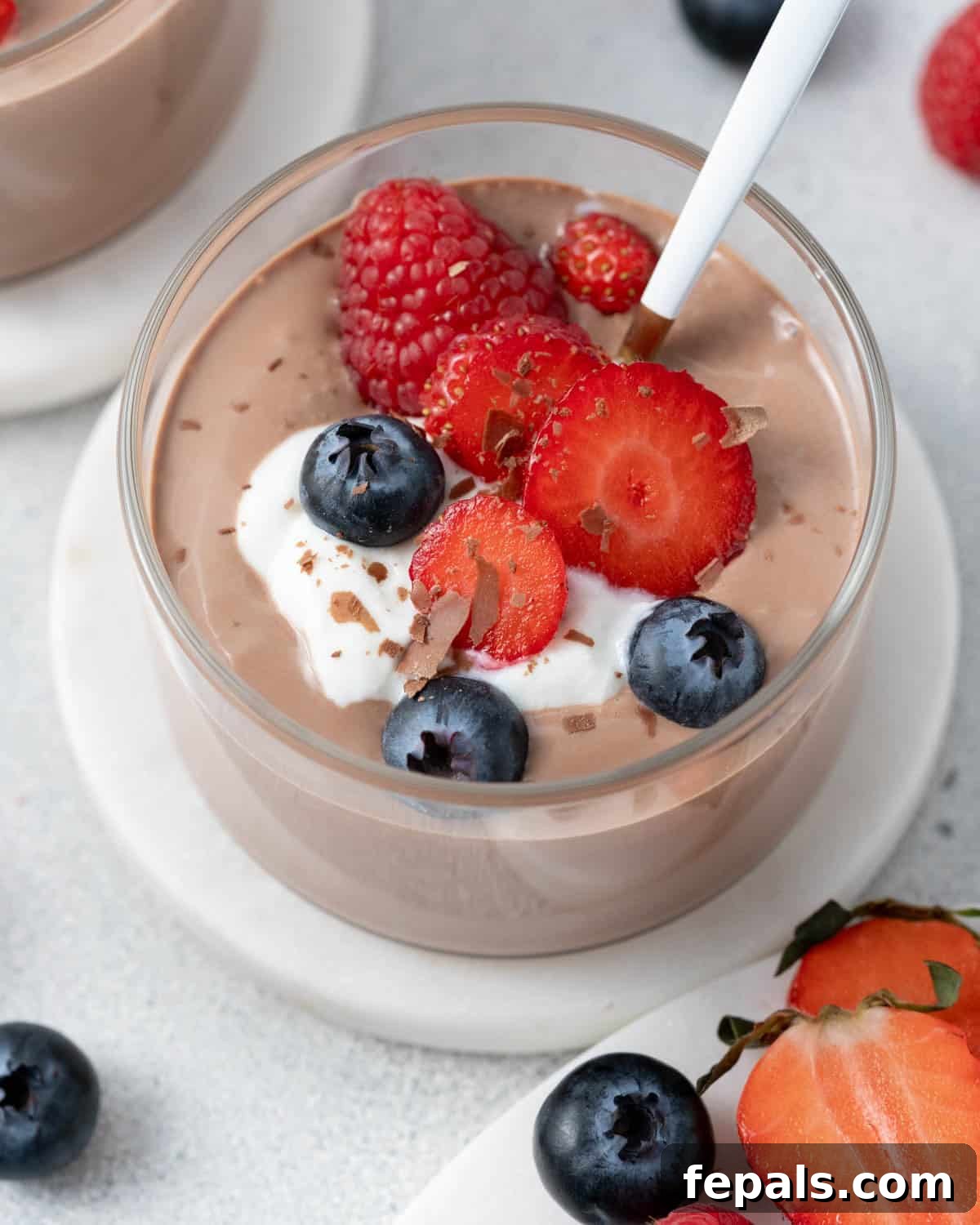 Close up of mousse in a glass cup with a spoon, topped with berries and set on a small white plate.