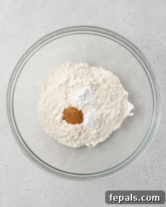 Flour, cinnamon, baking powder, baking soda, and salt, added to a bowl and ready to be mixed.