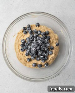 Blueberries coated in flour being added over muffin batter in a mixing bowl.