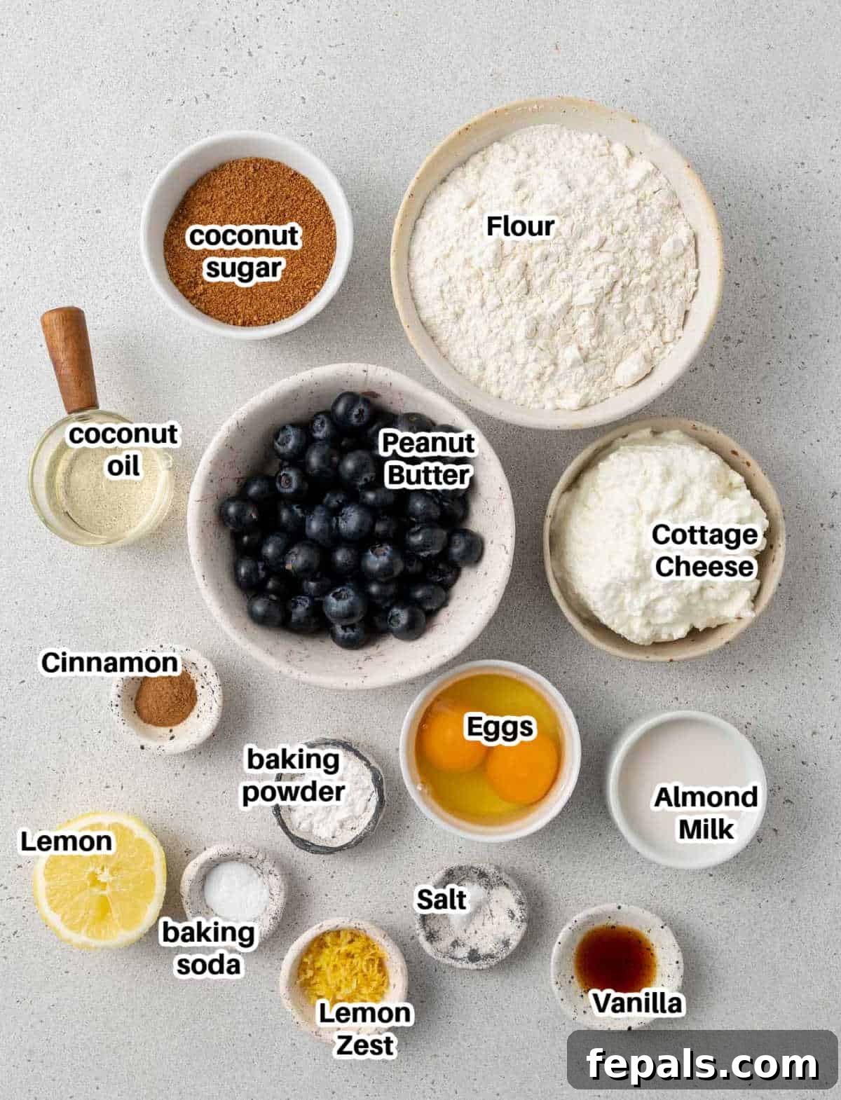 A collection of fresh ingredients laid out, including flour, blueberries, eggs, milk, cottage cheese, sugar, and baking essentials, ready to make blueberry muffins.