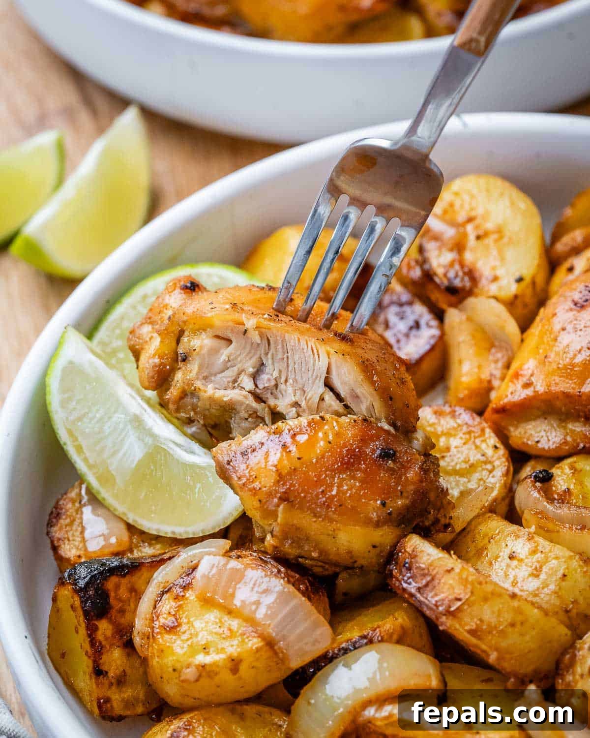 Fork cutting into a juicy chicken thigh over a bed of seasoned roasted potatoes with lime wedges nearby, ready to be squeezed.