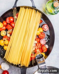 Spaghetti is added over tomatoes in a skillet with a water kettle pouring water over it.