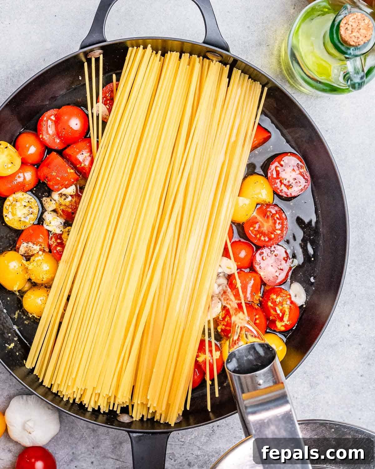 Spaghetti is added over tomatoes in a skillet with a water kettle pouring water over it.