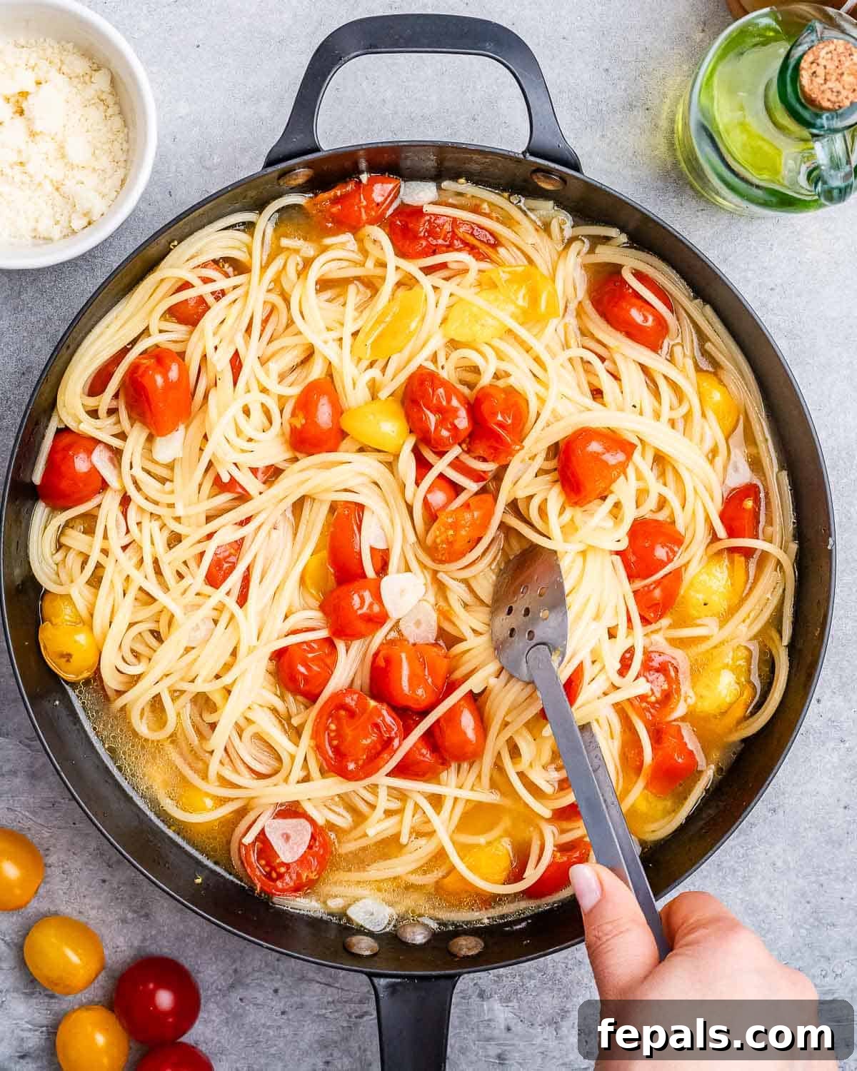 hand holding tongs and mixing the pasta and cherry tomatoes in a skillet.