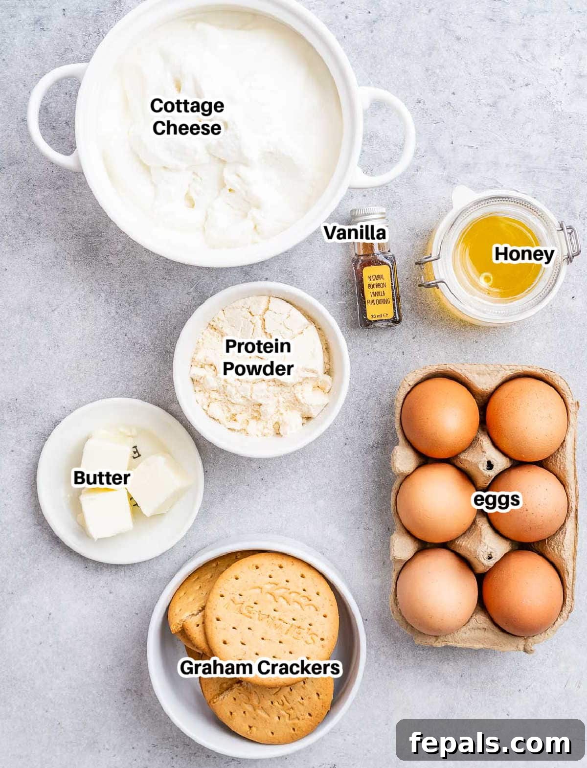 Top view of all ingredients for cottage cheese cheesecake neatly arranged in small bowls on a white surface: cottage cheese, eggs, graham crackers, honey, protein powder, butter, and vanilla extract.
