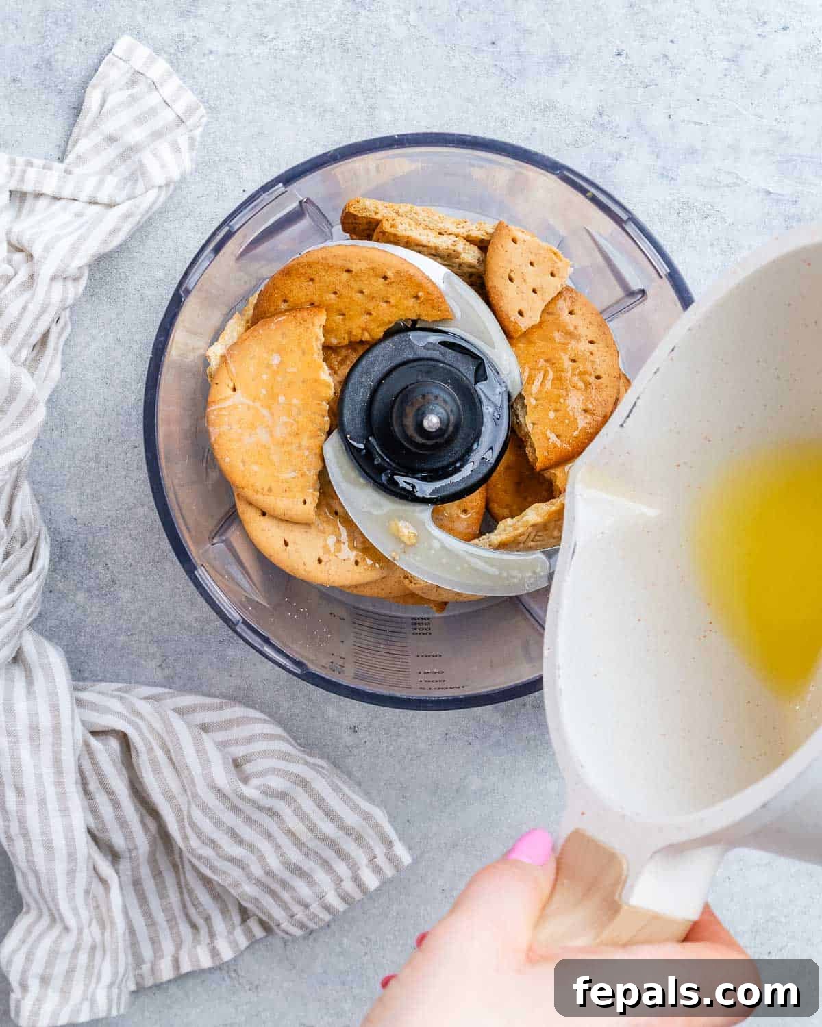 Melted butter being poured into a food processor with broken graham crackers, ready to be blended for the crust.