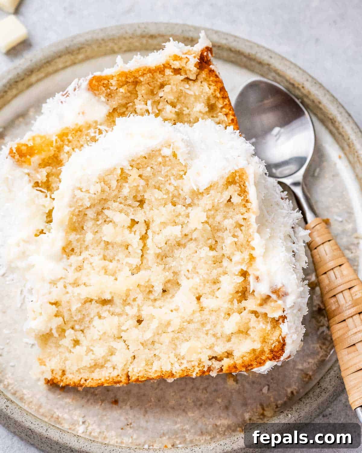 Tropical Coconut Bliss Cake 2 A generous slice of moist coconut cake on a beige plate, beautifully topped with fluffy cream cheese frosting and a sprinkle of shredded coconut. A silver spoon rests beside it, inviting a taste.