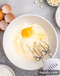 Tropical Coconut Bliss Cake 15 Egg being blended into the butter and sugar mixture using an electric mixer, with cracked eggs nearby.