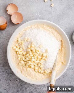Tropical Coconut Bliss Cake 17 White chocolate chunks and shredded coconut being folded into thick cake batter in a mixing bowl.