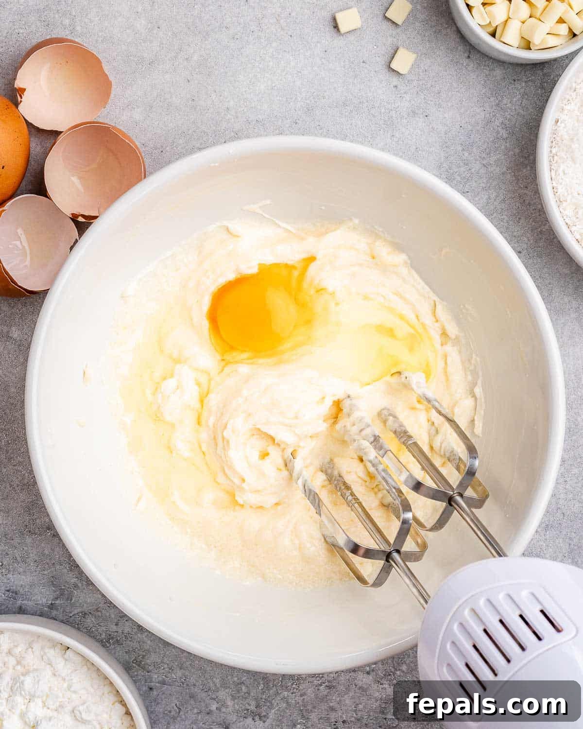 Tropical Coconut Bliss Cake 5 An egg being carefully blended into the creamy butter and sugar mixture using an electric mixer, with several cracked eggs waiting nearby for subsequent additions, ensuring each is fully incorporated.