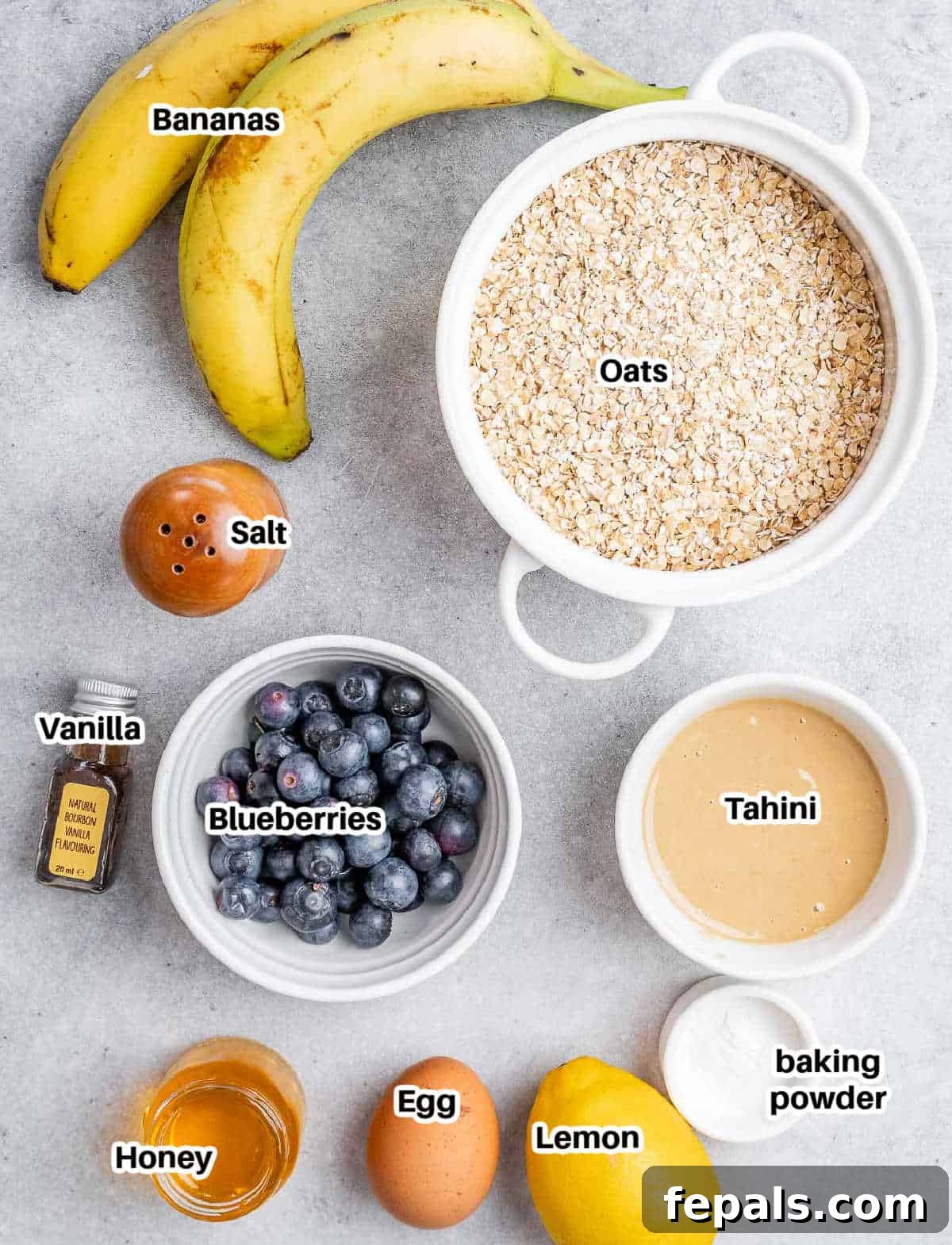 Soft-Baked Blueberry Oatmeal Cookies 3 A flat lay of ingredients laid out on a white surface, including bananas, honey, tahini, vanilla, an egg, lemon, quick oats, baking powder, salt, and fresh blueberries, ready to make blueberry oatmeal cookies.